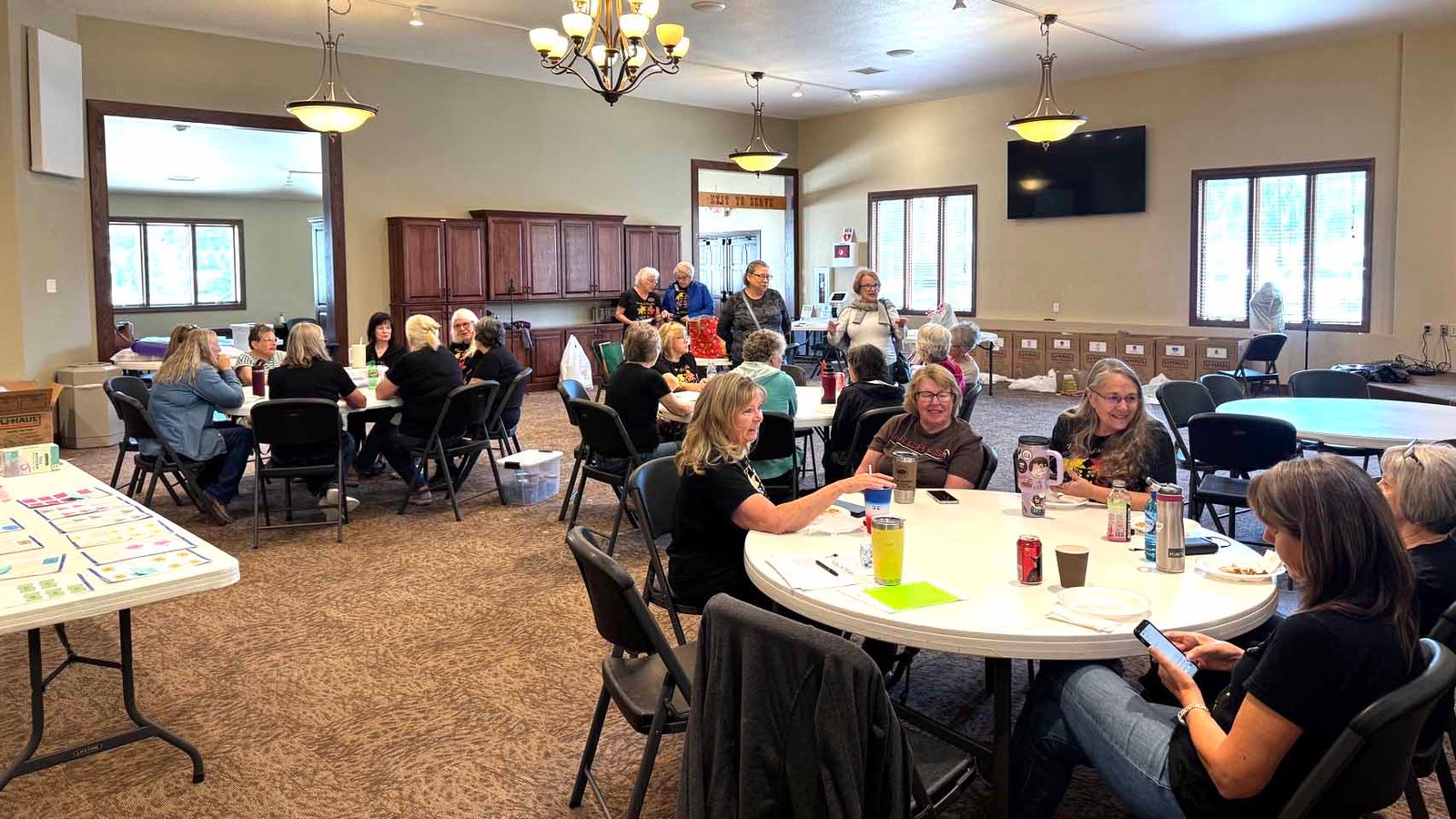A group of women from the Heart of the Hills Quilters Guild enjoying a relaxed break during quilt intake for the 2025 Hill City Quilt Show. Members are seated at round tables chatting and socializing in a bright community hall.